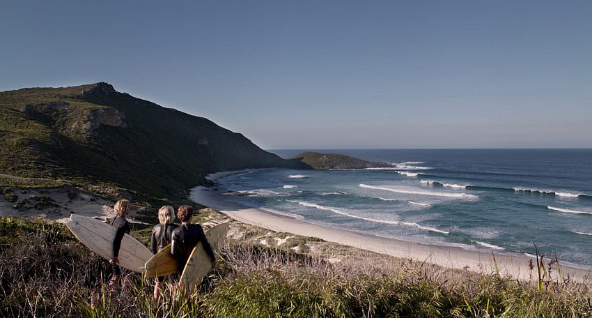 Three people holding surfboards looking out at a sandy cove with waves crashing against the beach