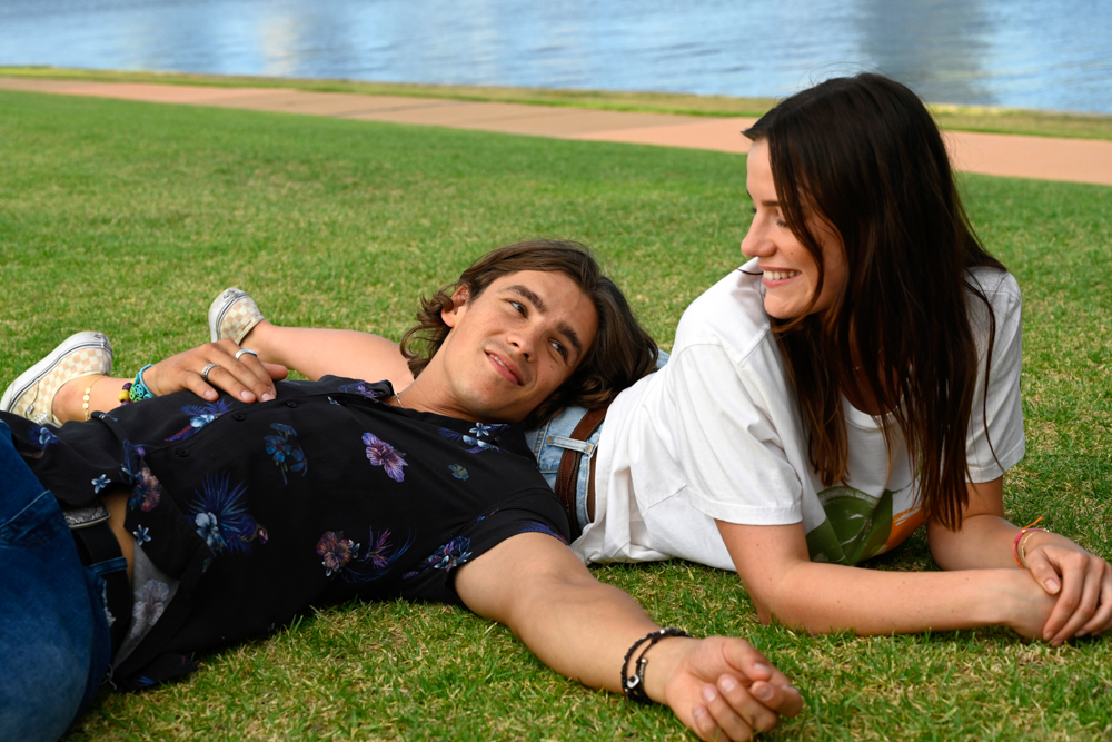 Lily Sullivan (as Lucy) & Brenton Thwaites (as Devon) lying on grass by the river.