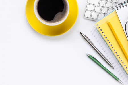 White desk with a yellow cup and saucer filled with black coffee next to a keyboard, yellow notebook, reading glasses and pens in the top right-hand corner.